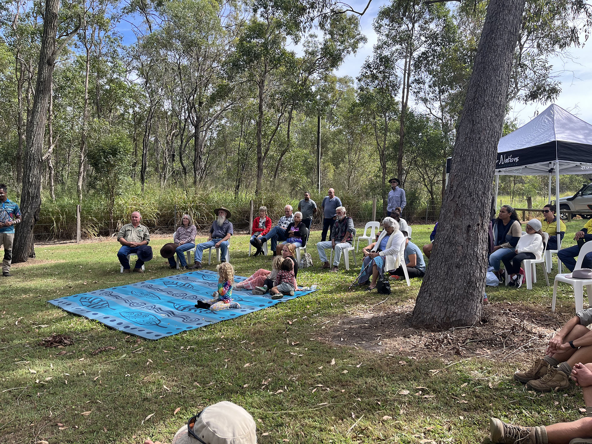 A group of people sit on fold-up chairs and blankets in a cleared area near native vegetation